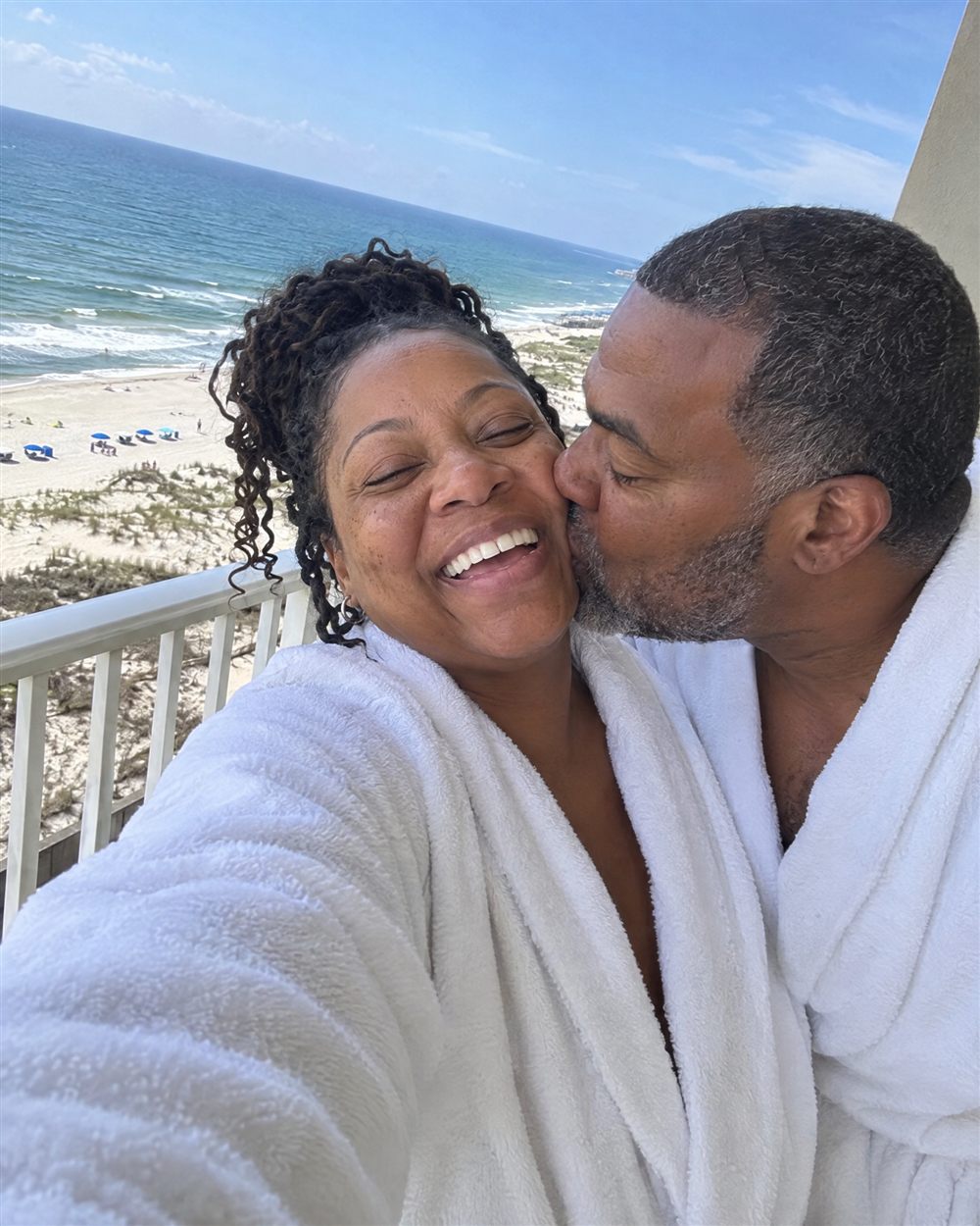 Couple in white bathrobes taking a selfie on a balcony above the beach
